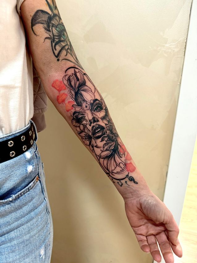 Tatuaje blackwork de un rostro femenino entre flores con manchas rojas en acuarela, en el antebrazo