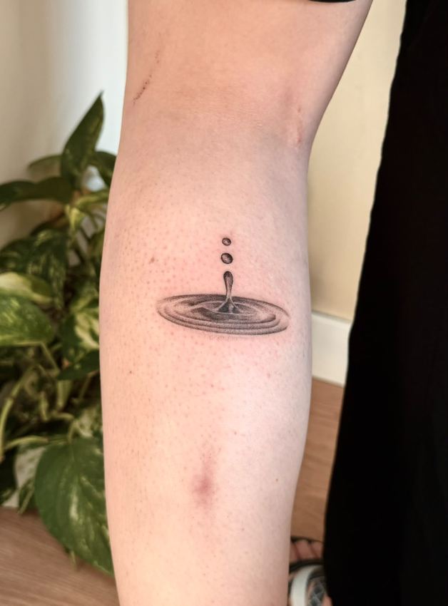 Tatuaje de microrealismo de una gota de agua cayendo sobre una superficie, en el brazo